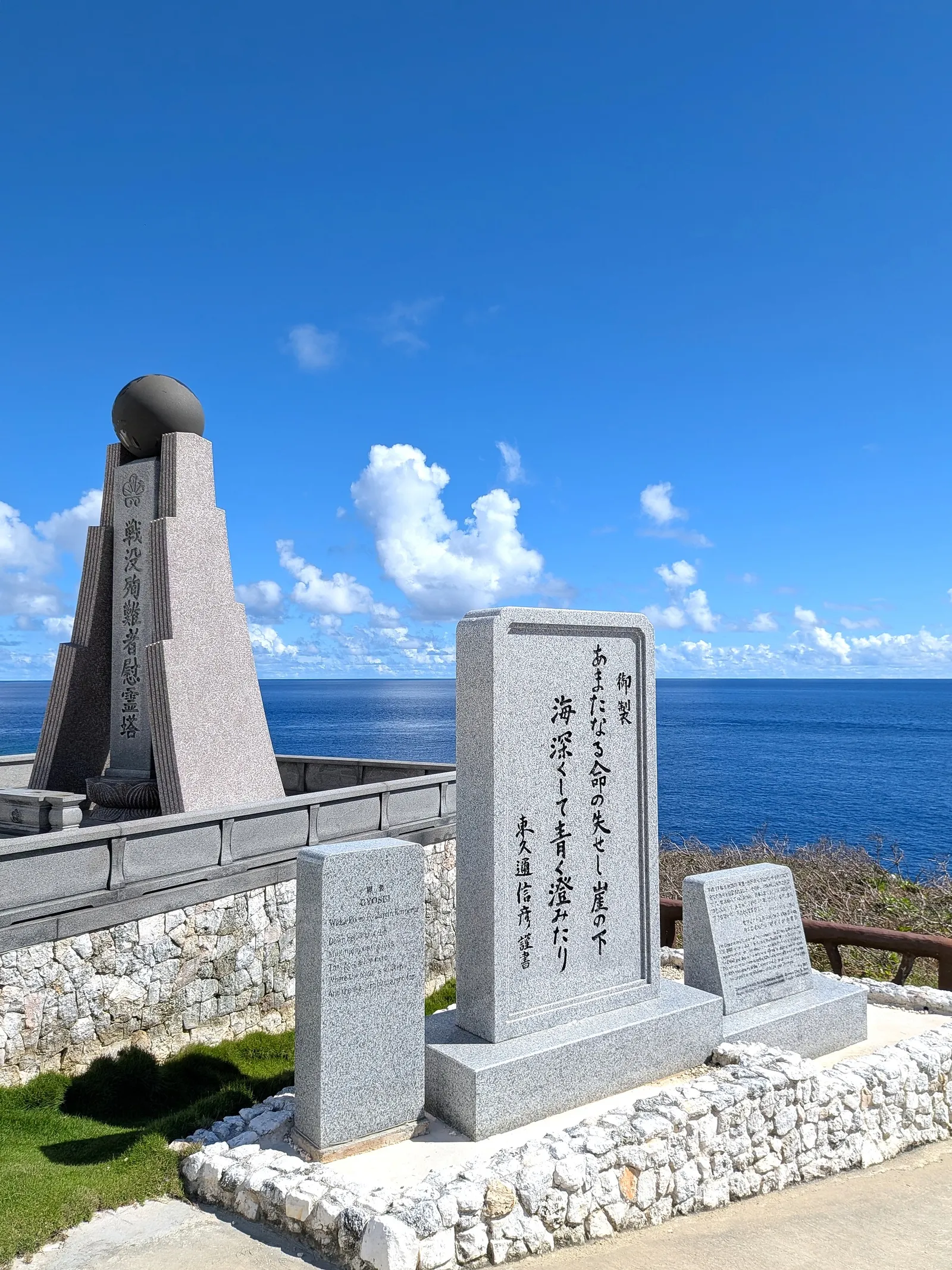 A pale granite stone at Banzai Cliff inscribed with a Japanese poem, standing beside the dark memorial tower above the sea
