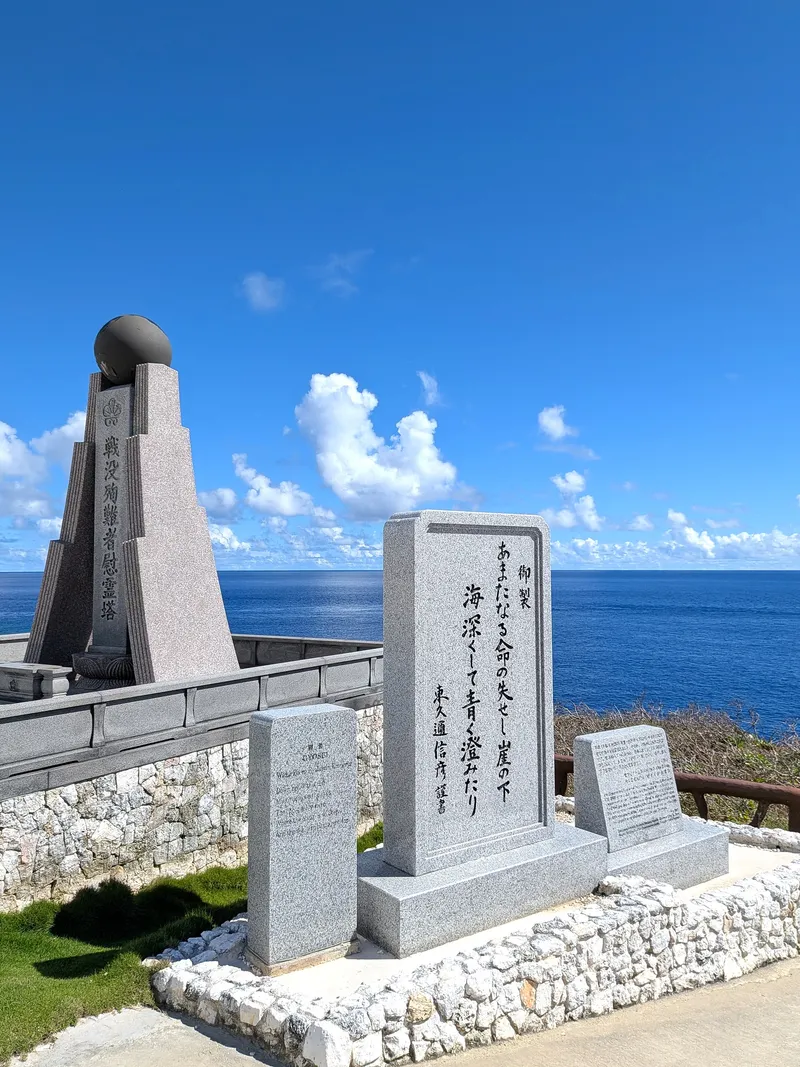 A pale granite stone at Banzai Cliff inscribed with a Japanese poem, standing beside the dark memorial tower above the sea