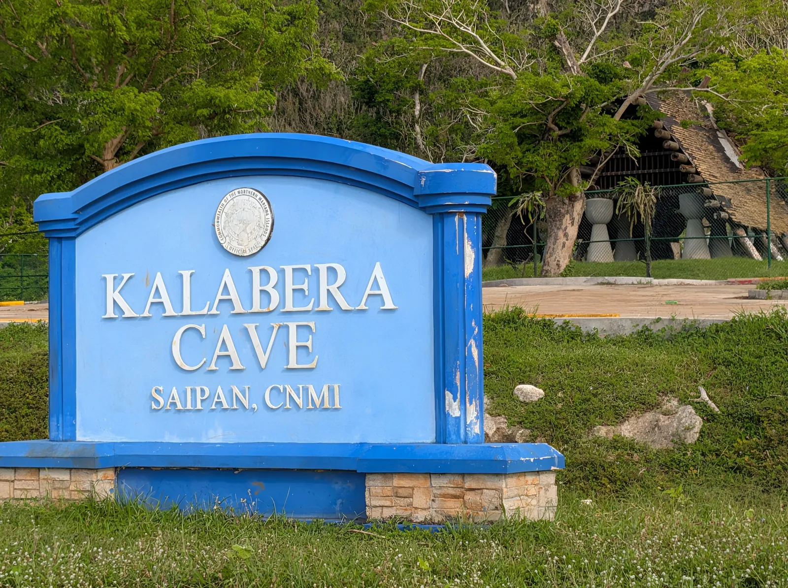 The blue Kalabera Cave entrance sign carrying the CNMI seal, with the latte hut visible behind it