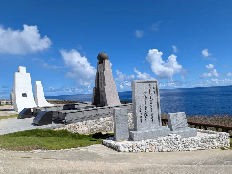 The main cluster of monuments at Banzai Cliff, Saipan — a dark granite memorial tower, an inscribed imperial poem stone, and the white pylons of the Tower of the Pacific, with open ocean behind