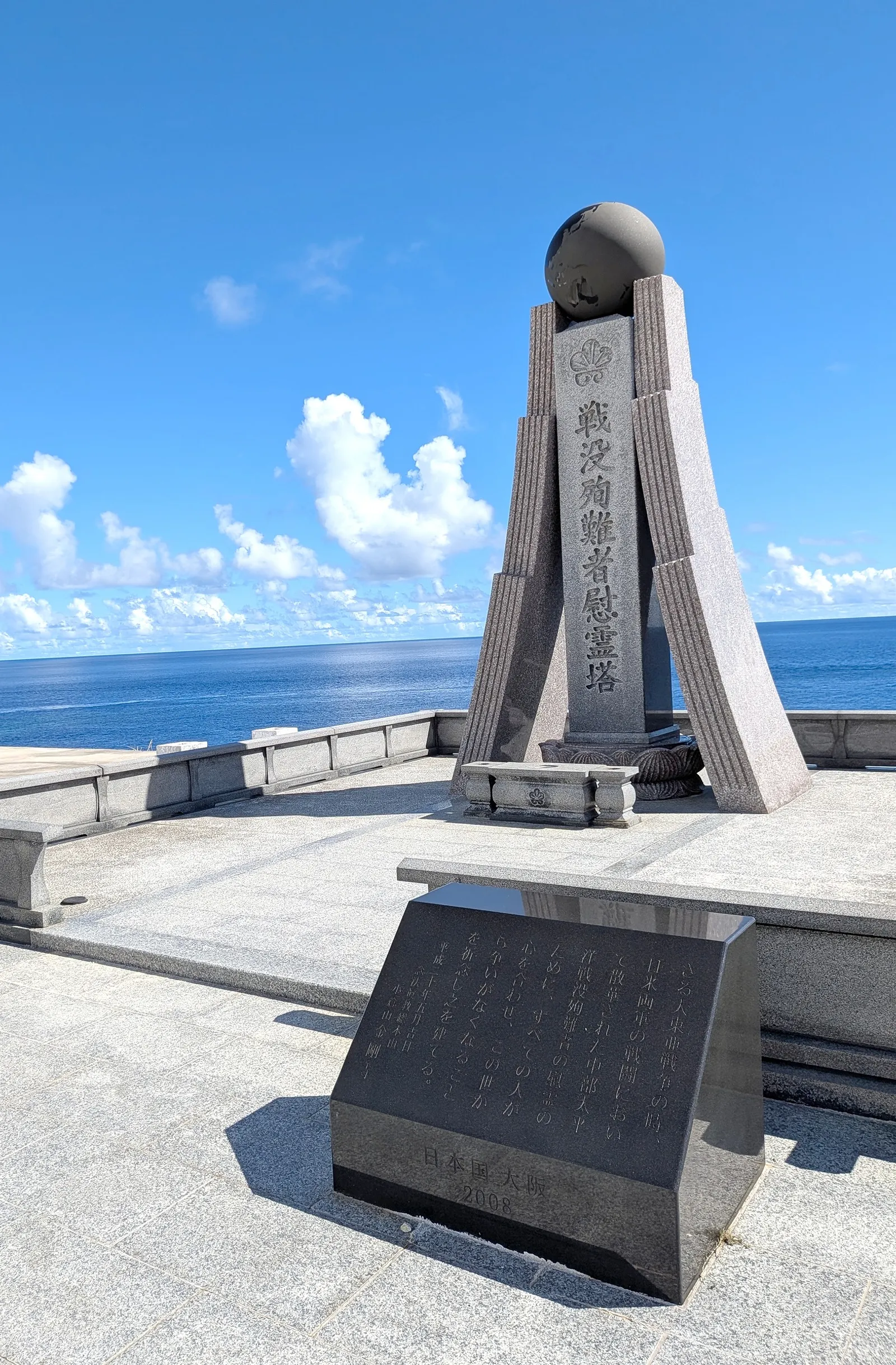 The dark granite memorial tower at Banzai Cliff, topped with a stone sphere and bearing a chrysanthemum crest, with an inscribed plaque placed in front of it