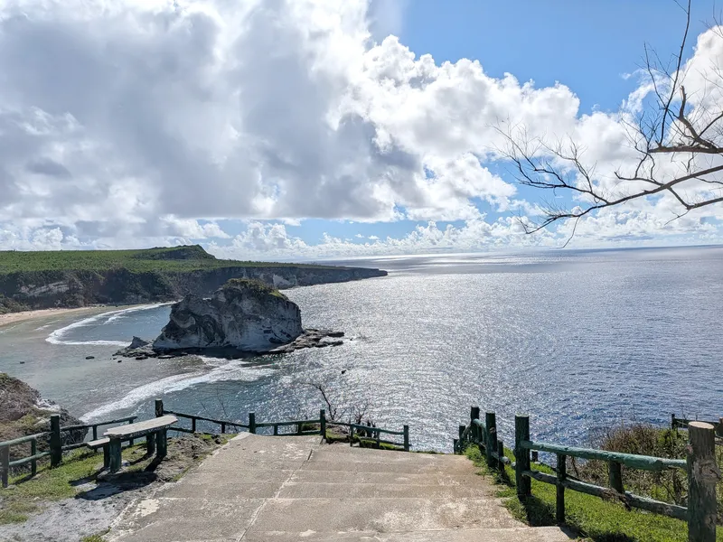 The view from Bird Island Lookout, Saipan — a limestone islet in a turquoise bay, with the stepped concrete path and bench seating of the overlook in the foreground