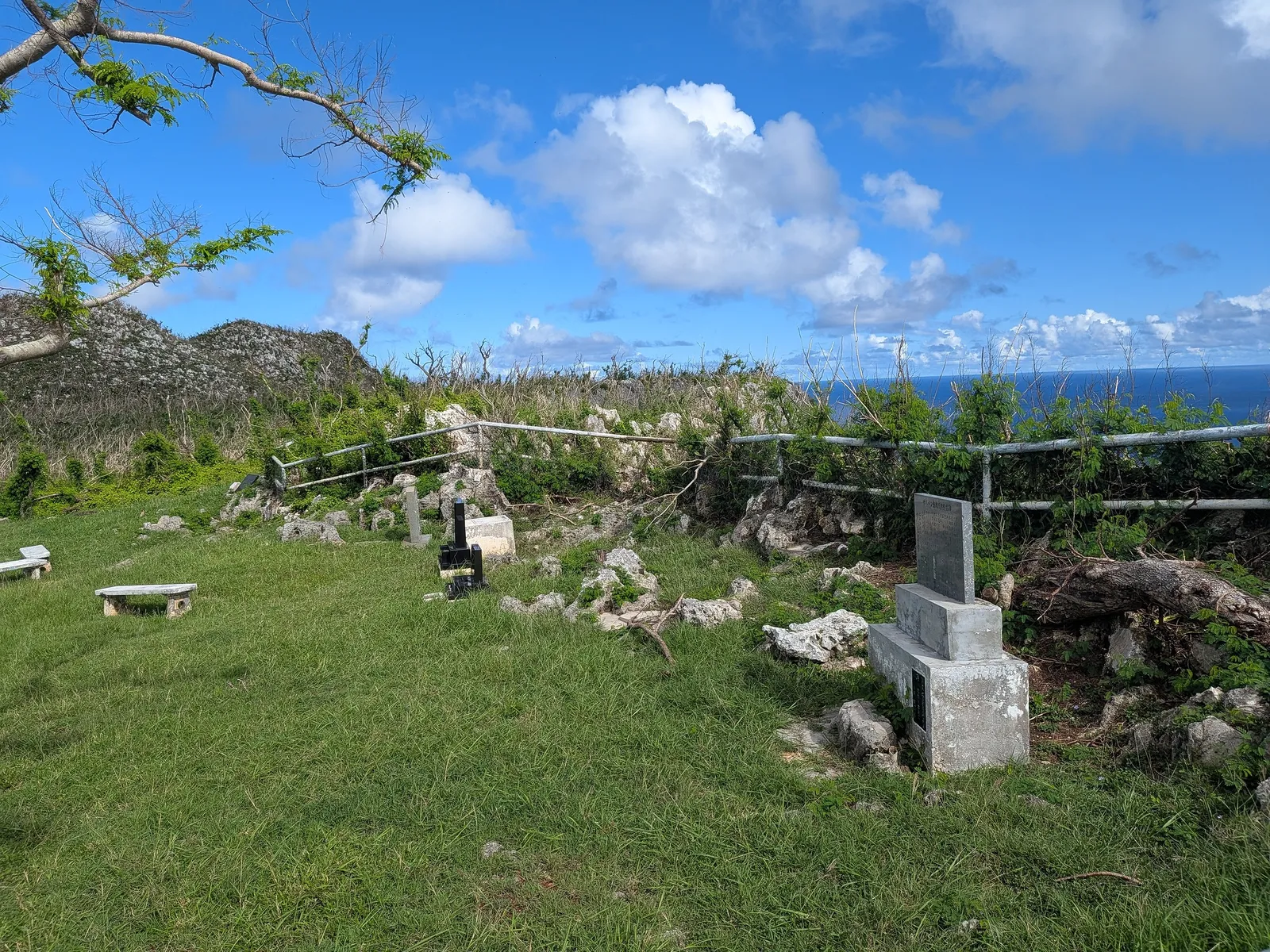 Smaller cenotaphs and grave markers on the grass at Suicide Cliff, with the ocean visible beyond the clifftop railing