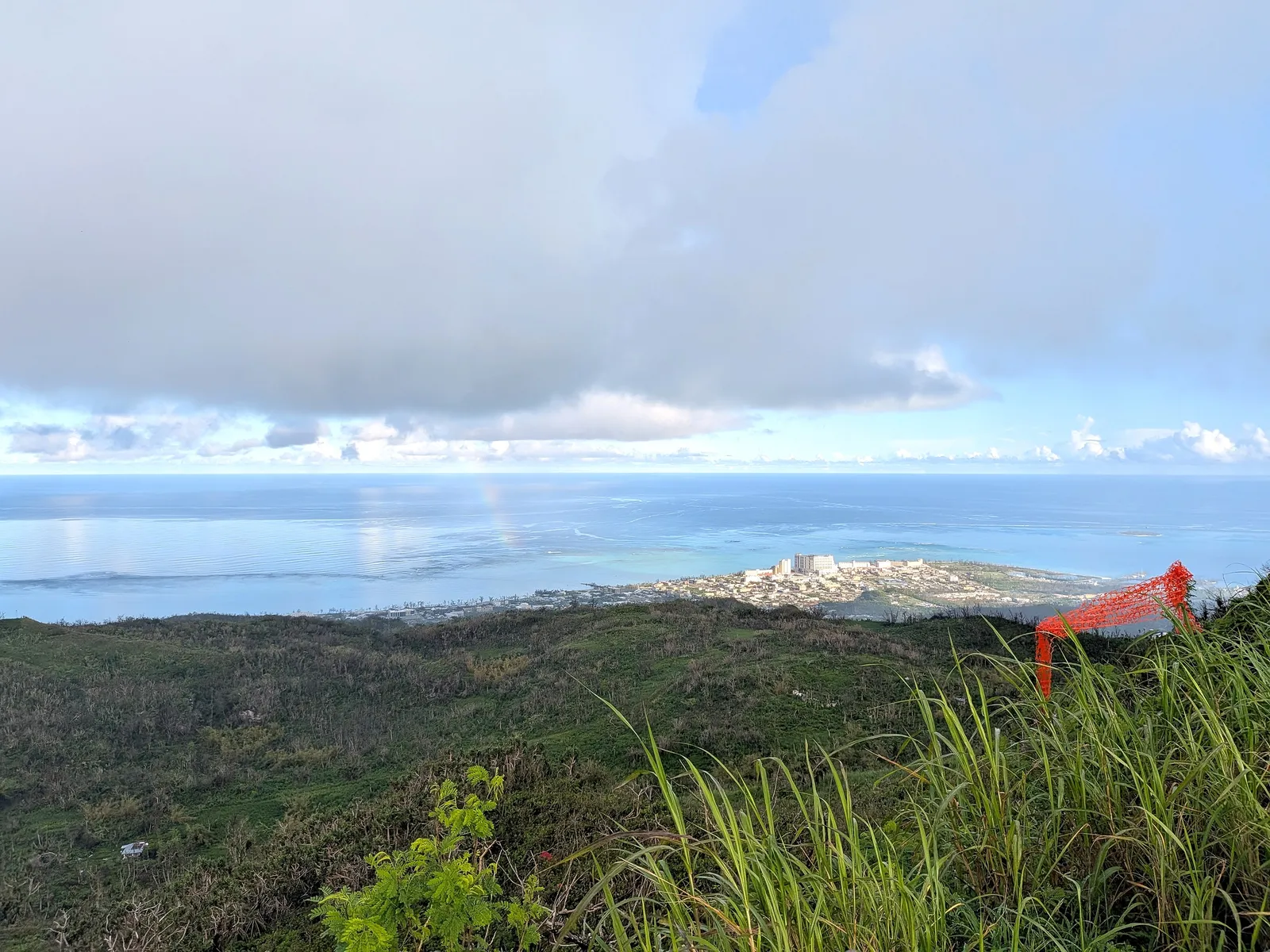 A wide view from Mount Tapochau's lower slopes with a faint rainbow over the coastal plain